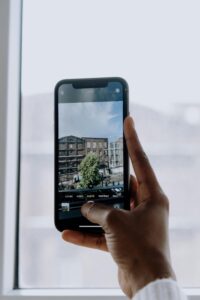 Mano sosteniendo un teléfono móvil que está tomando una fotografía de una calle con un edificio y un árbol.