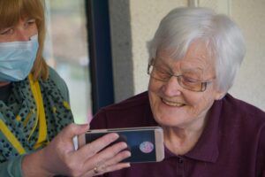 La imagen muestra a una mujer con mascarilla enseñando la pantalla de un smartphone a otra mujer de mayor edad, es una buena representación de la relación de algunas personas mayores y la tecnología y cómo existen dependencias para su manejo diario.