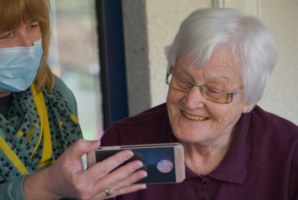 La imagen muestra a una mujer con mascarilla enseñando la pantalla de un smartphone a otra mujer de mayor edad, es una buena representación de la relación de algunas personas mayores y la tecnología y cómo existen dependencias para su manejo diario.